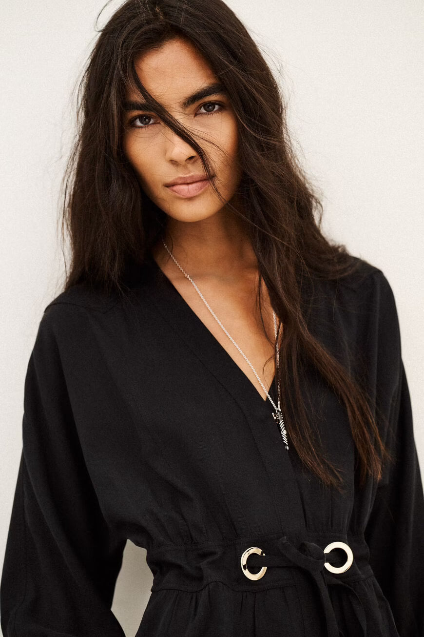 A woman with long dark hair wearing a black wrap-style top with a tie waist and silver-tone ring details, photographed against a plain light background.