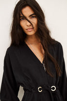 A woman with long dark hair wearing a black wrap-style top with a tie waist and silver-tone ring details, photographed against a plain light background.