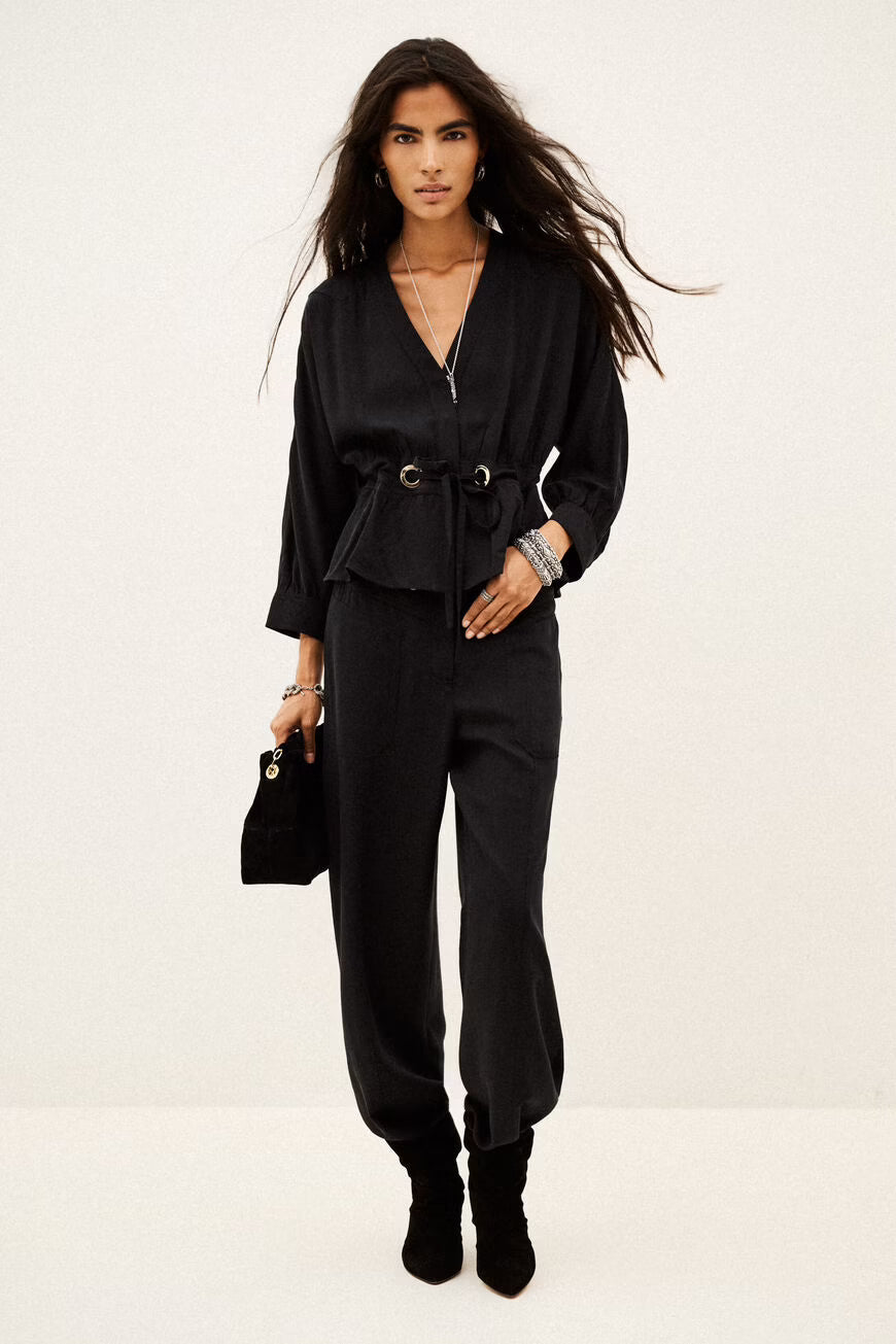 A model wearing a black long-sleeve belted top and matching black pants, holding a small black handbag against a plain off-white studio background.