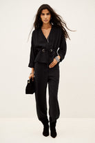A model wearing a black long-sleeve belted top and matching black pants, holding a small black handbag against a plain off-white studio background.