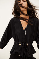 A woman wearing a black belted blouse with gold-tone ring details at the waist, styled with a silver pendant necklace. Her dark hair is blowing in the wind against a plain white background.