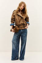 A woman modeling a brown double-breasted trench jacket with rolled-up sleeves, paired with wide-leg blue jeans and a matching brown belt bag, against a plain off-white studio background.