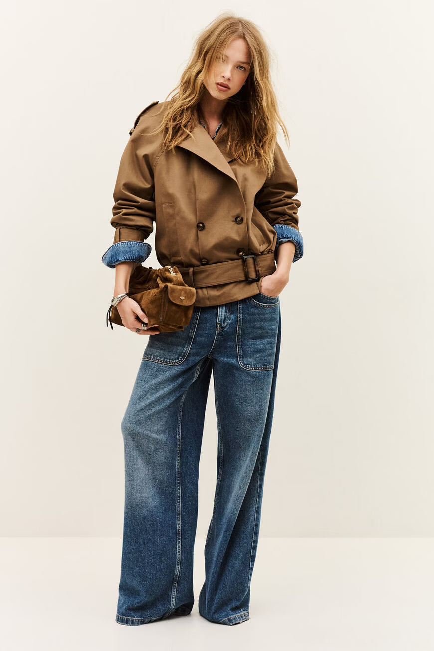A woman modeling a brown double-breasted trench jacket with rolled-up sleeves, paired with wide-leg blue jeans and a matching brown belt bag, against a plain off-white studio background.