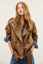 A woman wearing a brown double-breasted belted trench coat over a blue denim shirt and jeans, posing against a light off-white background.