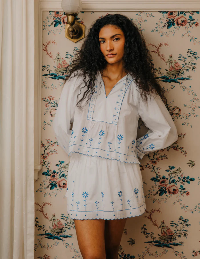 Woman wearing a white embroidered dress standing in front of floral wallpaper.
