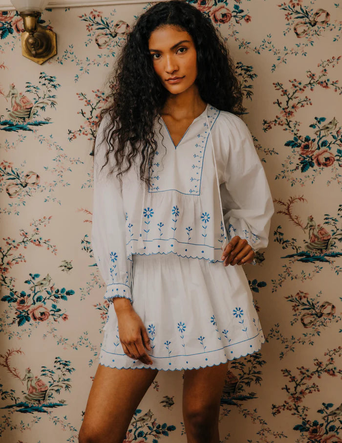 Woman in a white embroidered top and skirt standing against floral wallpaper.
