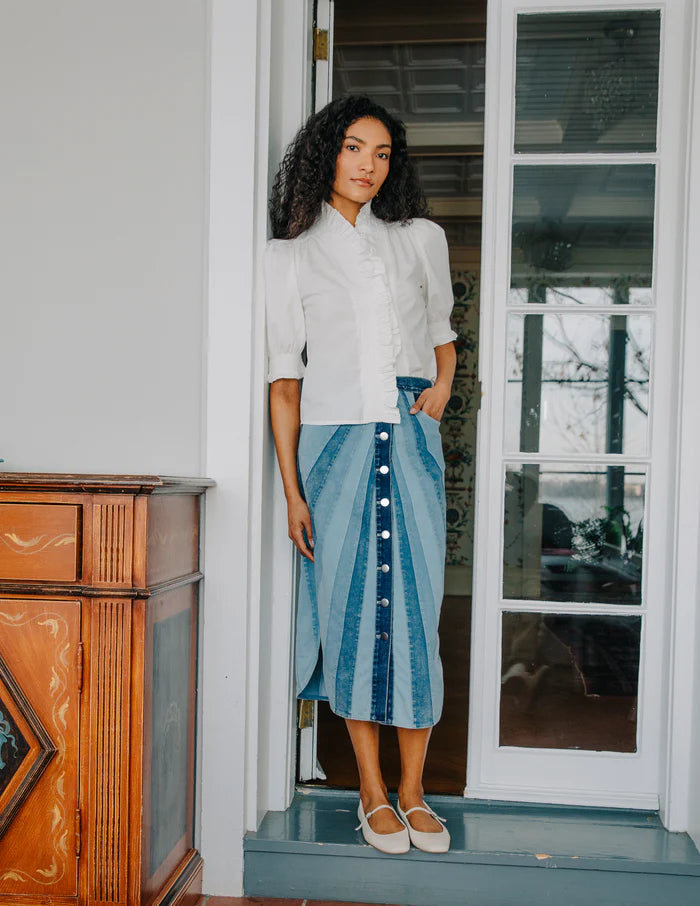 Woman standing on a porch wearing a white blouse and blue denim skirt.