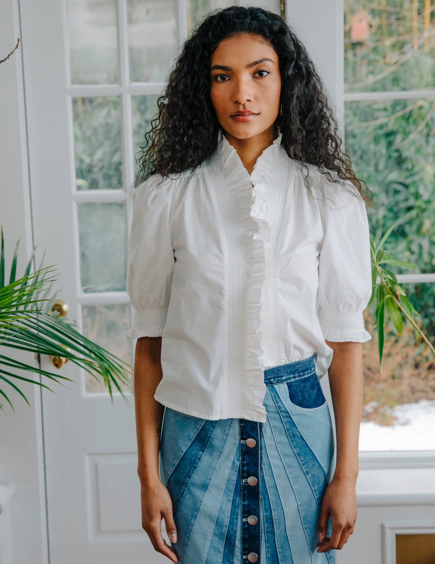Woman wearing a white blouse and blue denim skirt standing indoors with plants in the background