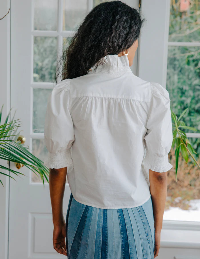 Person wearing a white blouse and blue skirt standing in a room with plants and a window.