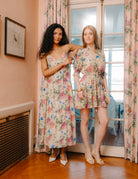 Two women wearing floral dresses standing in a room with floral curtains.