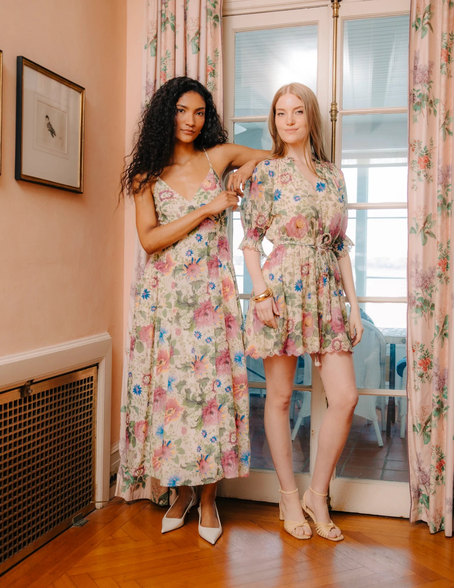 Two women wearing floral dresses standing in a room with floral curtains.