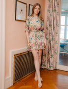 Woman in a floral dress standing in a room with pink walls and floral curtains.