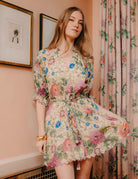 Woman wearing a floral dress standing in a room with framed pictures and curtains.