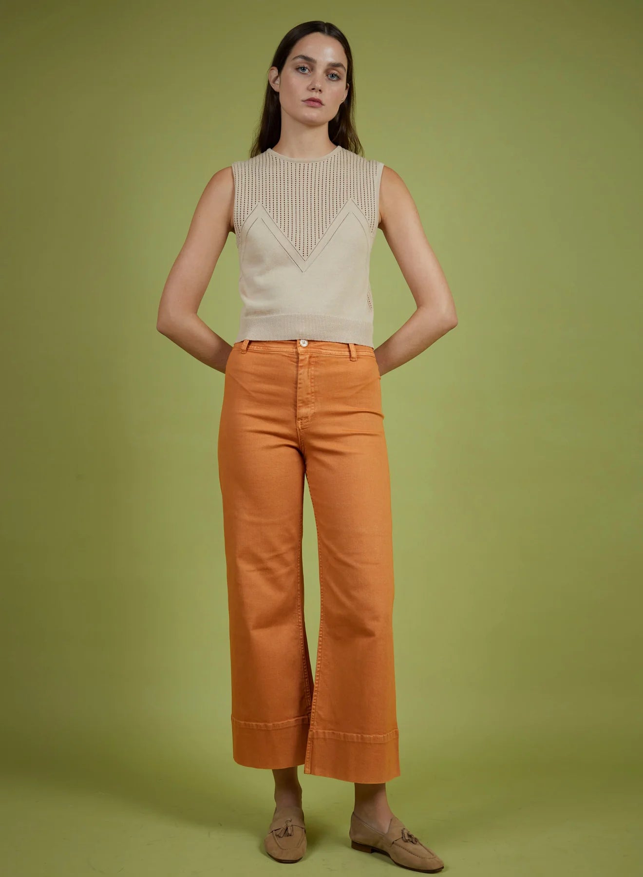 Woman wearing a beige sleeveless top and orange pants against a green background