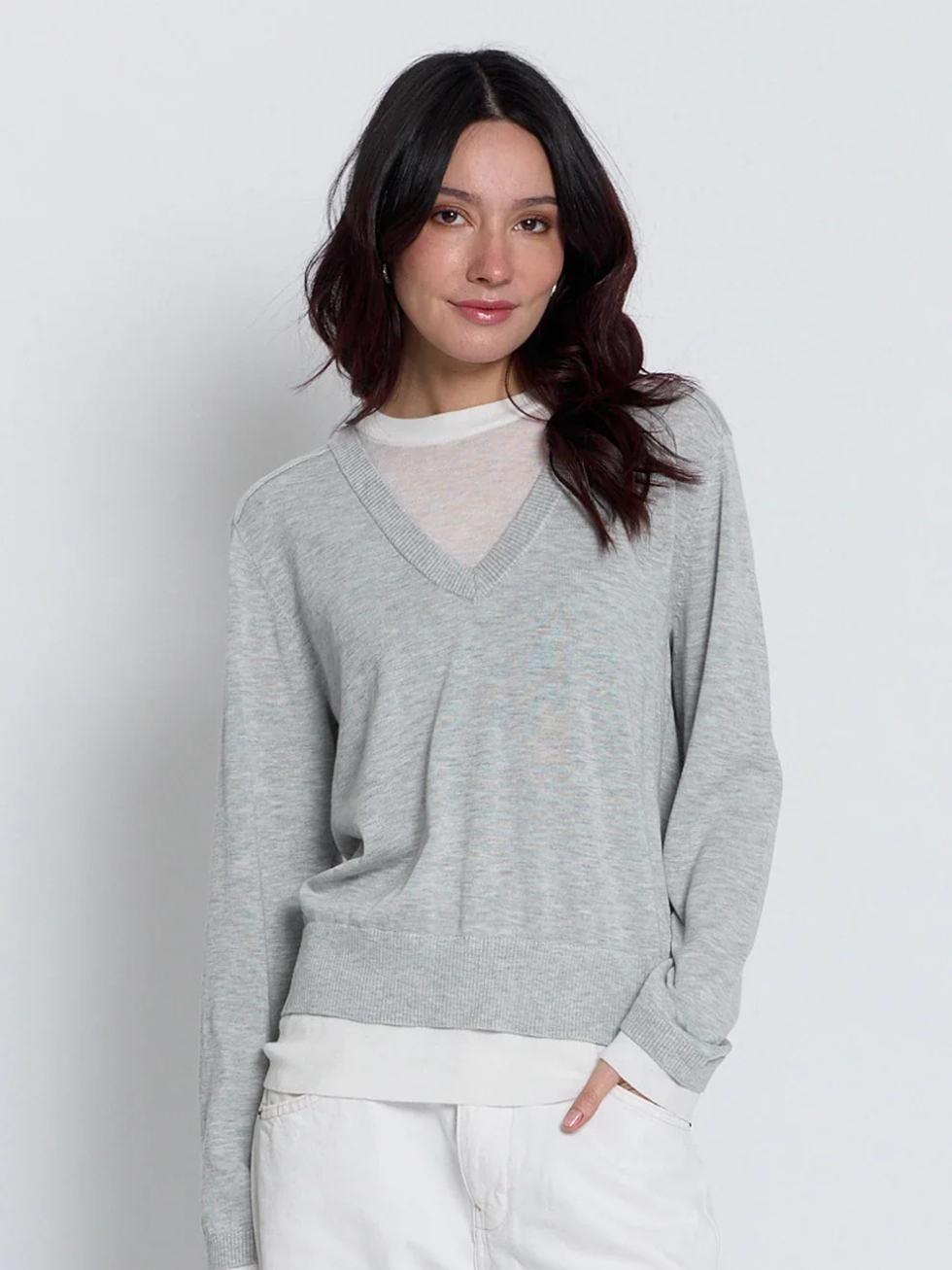 A woman wearing a light gray heathered V-neck knit sweater over a white mock neck top and white pants, standing against a light gray studio background.