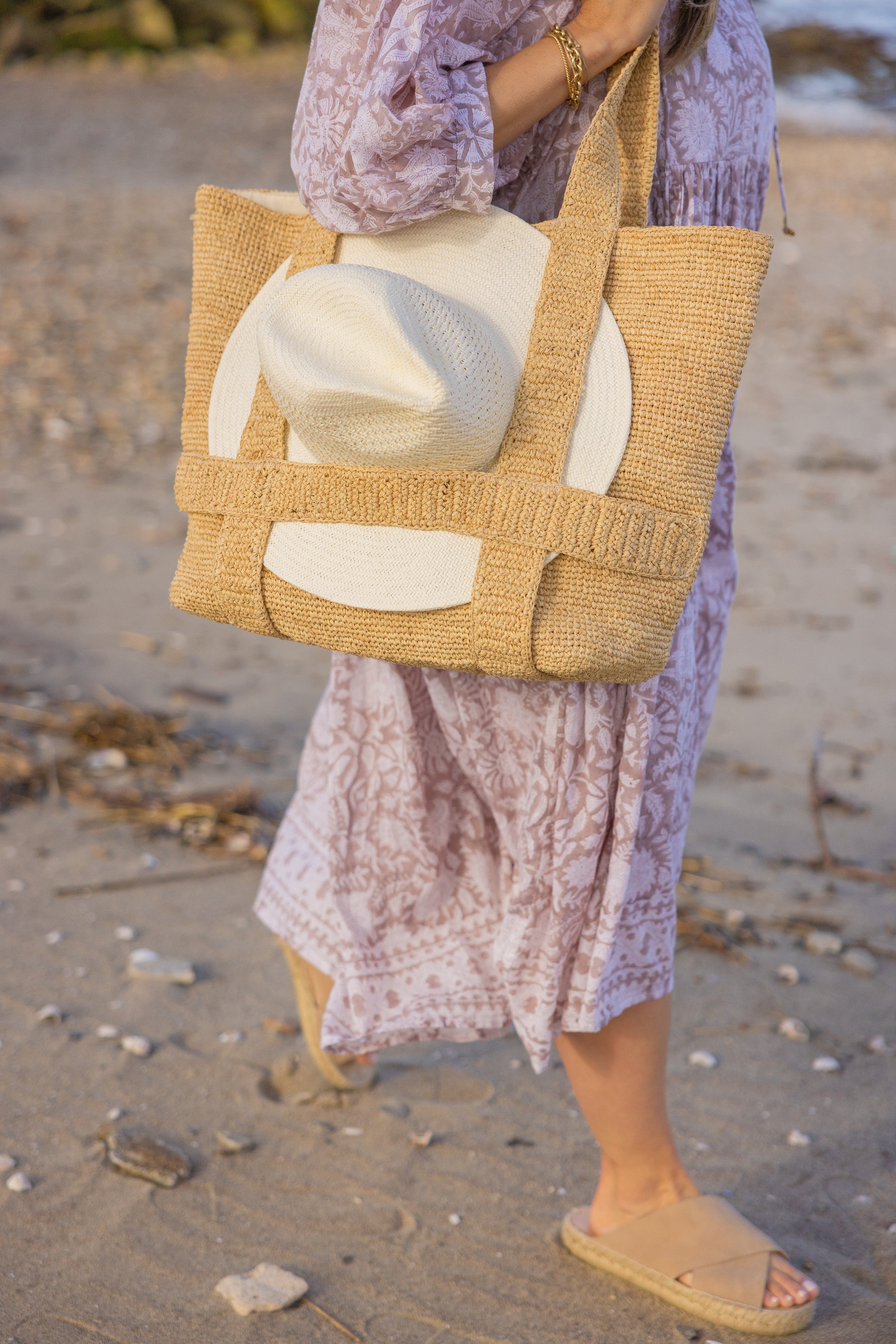 Person holding a woven bag with a white hat on a sandy beach