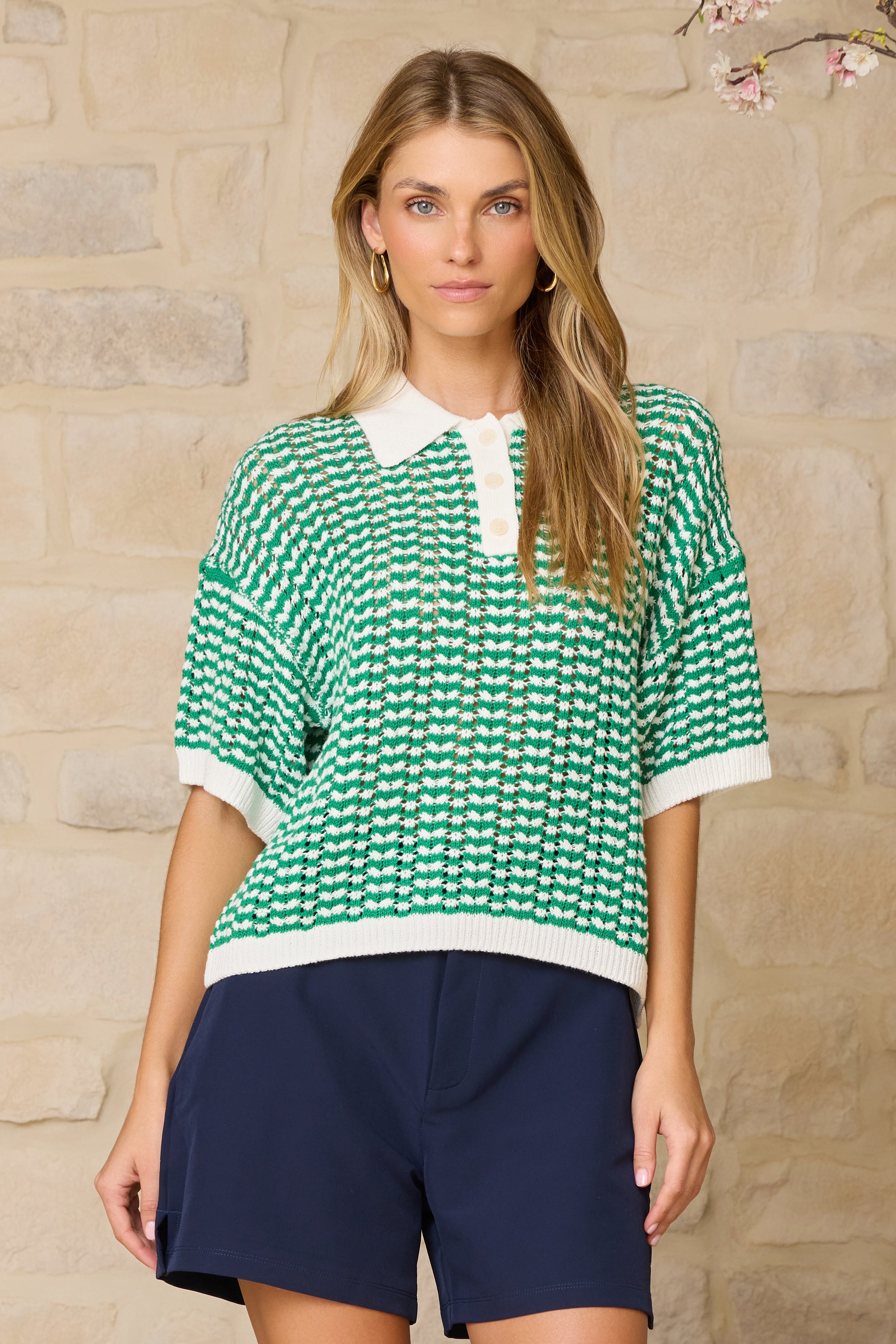 Woman wearing a green and white patterned shirt with navy shorts against a stone wall.
