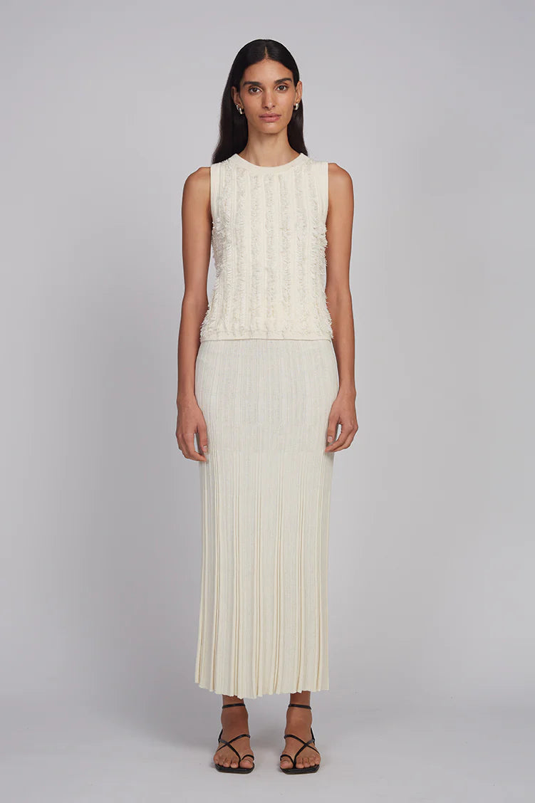 Woman wearing a sleeveless cream top and pleated skirt on a plain background