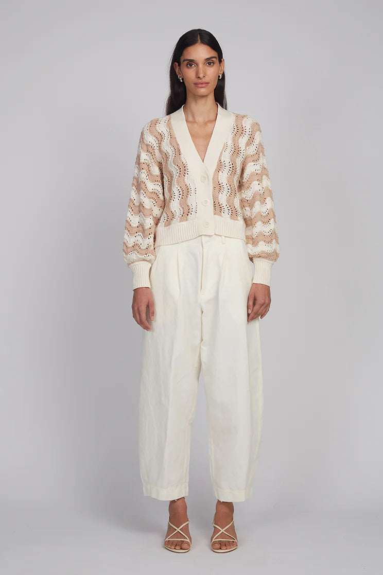 Woman wearing a patterned cardigan and white pants on a gray background