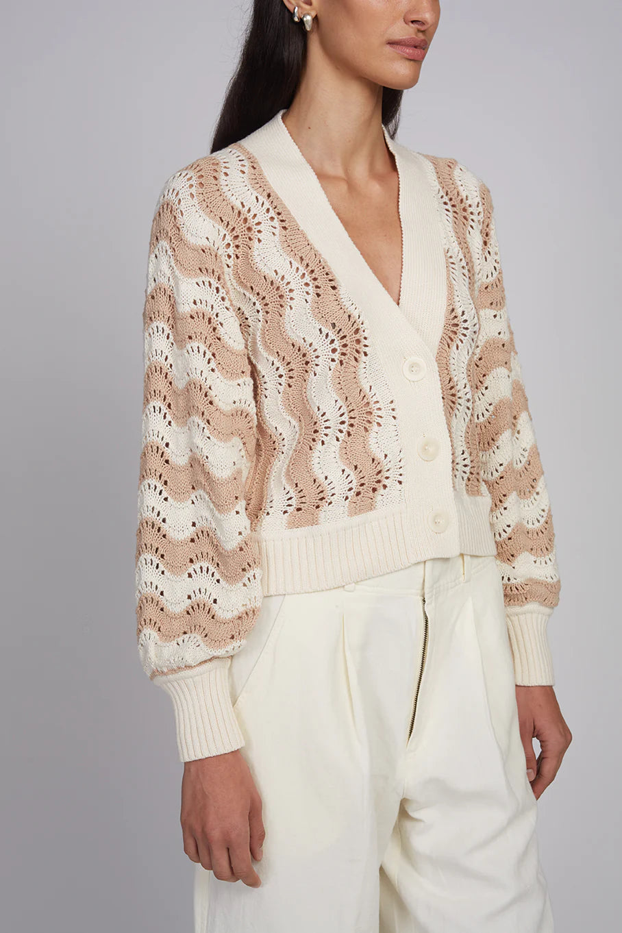 Woman wearing a beige and brown patterned sweater with white pants on a gray background