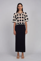Woman wearing a black and white polka dot top with a long black skirt on a gray background