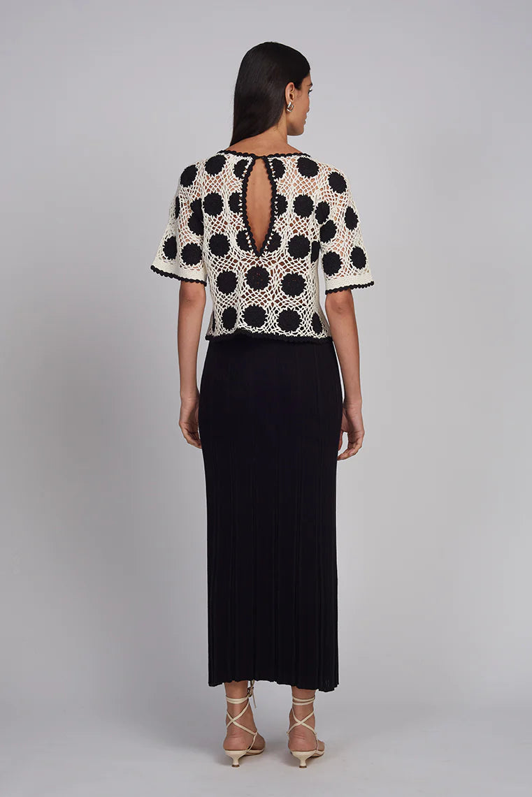 Woman wearing a black and white polka dot top with a cutout back design, paired with a black skirt on a gray background.