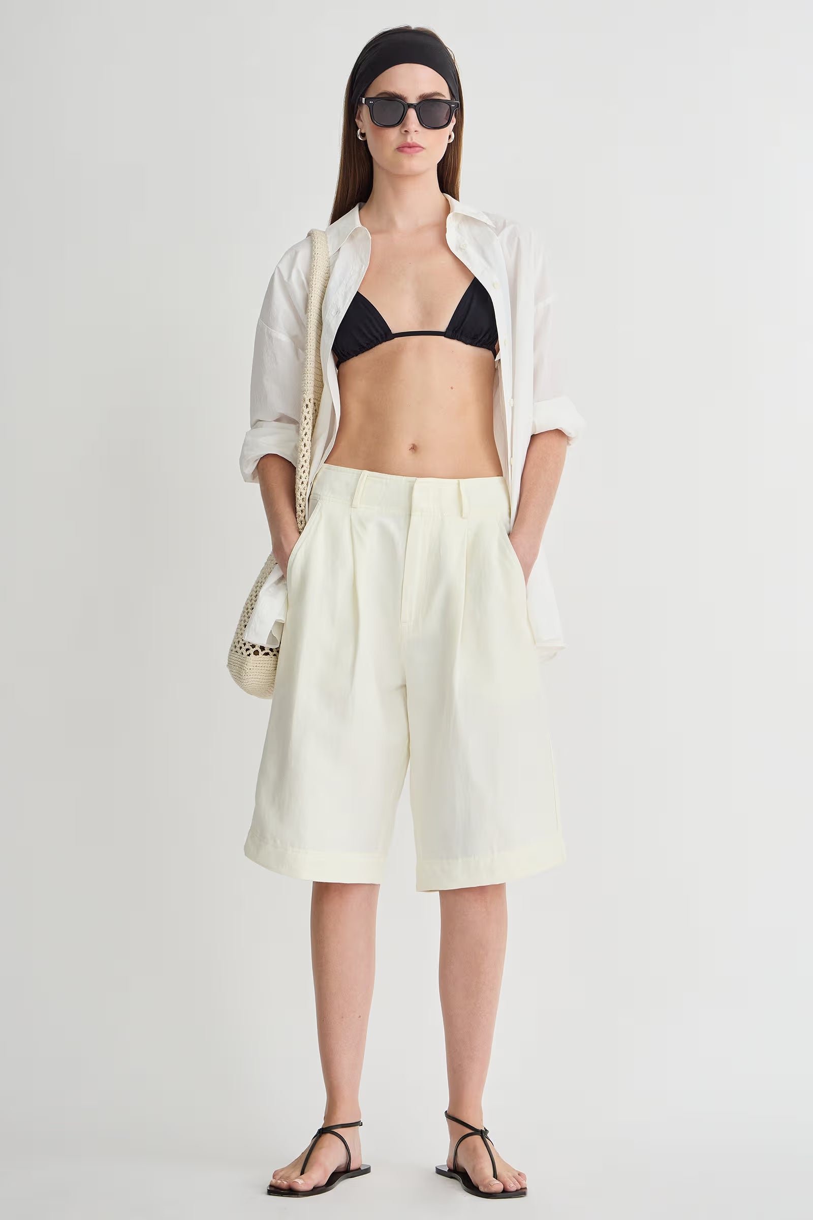 Woman wearing a black bikini top, white shirt, light yellow shorts, and black sandals on a white background.