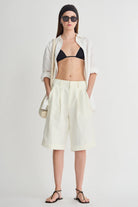 Woman wearing a black bikini top, white shirt, light yellow shorts, and black sandals on a white background.