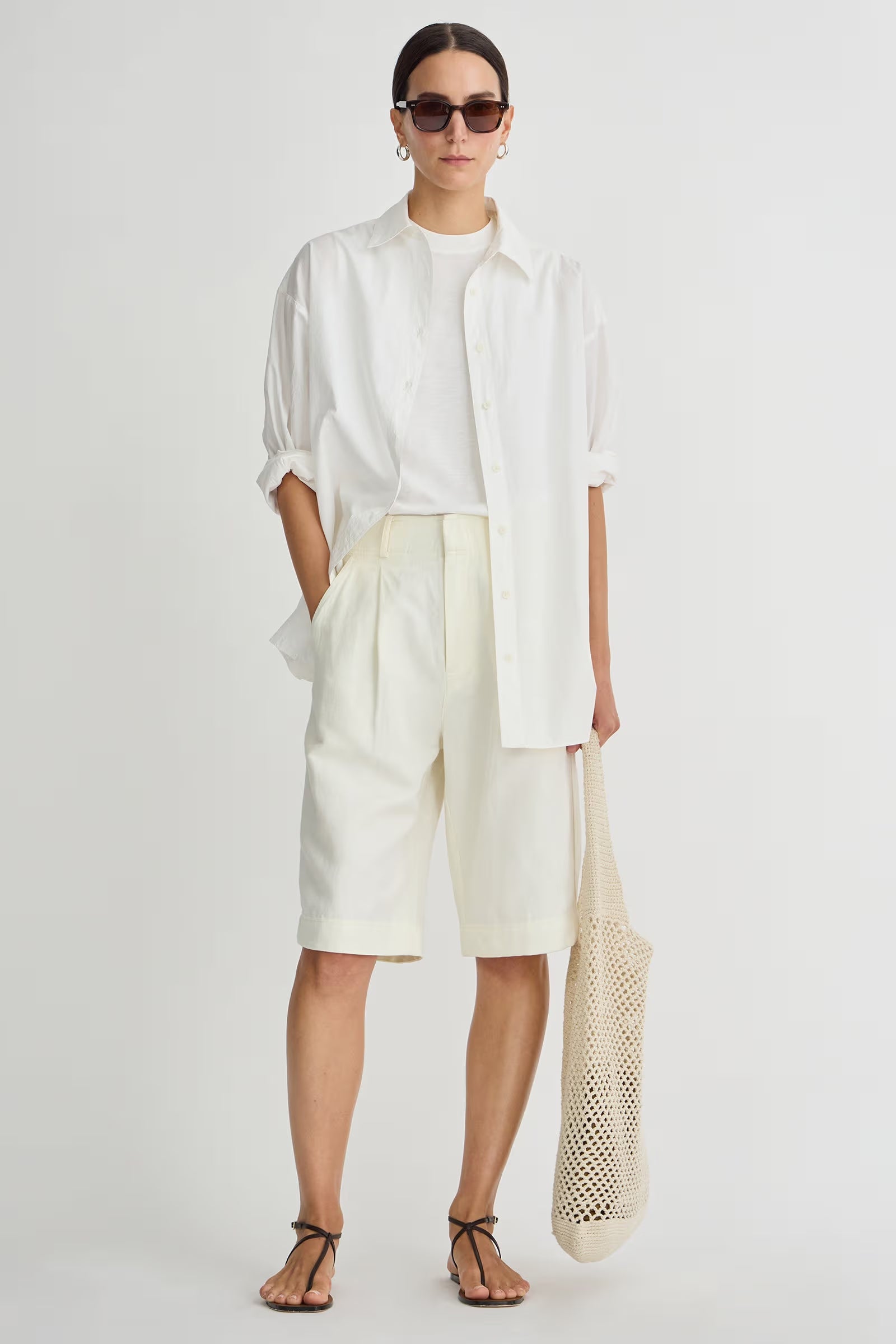 Person wearing a white outfit with sunglasses and holding a beige bag on a plain background