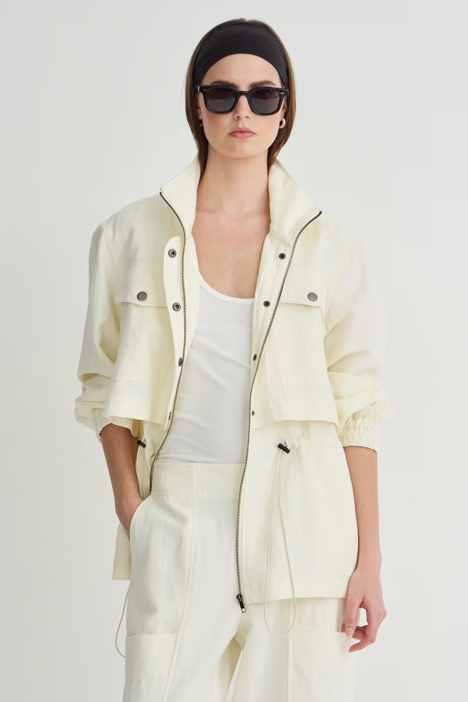 Woman wearing a light-colored jacket, white top, and pants on a plain background