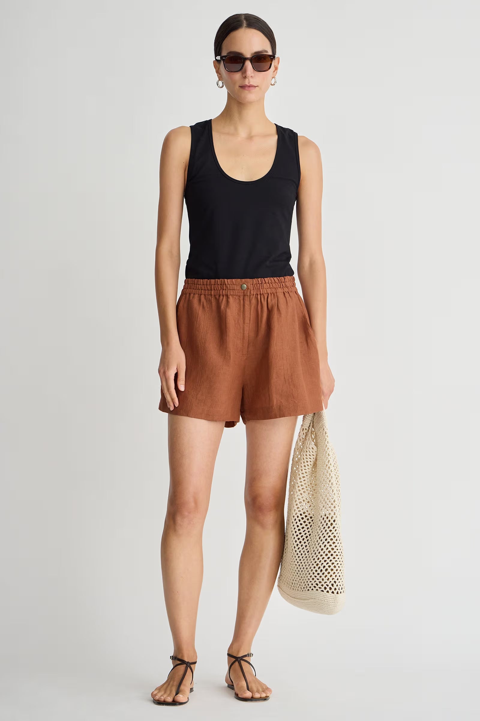 Woman wearing a black tank top and brown shorts, holding a beige mesh bag on a white background