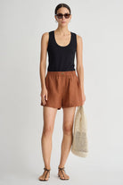 Woman wearing a black tank top and brown shorts, holding a beige mesh bag on a white background