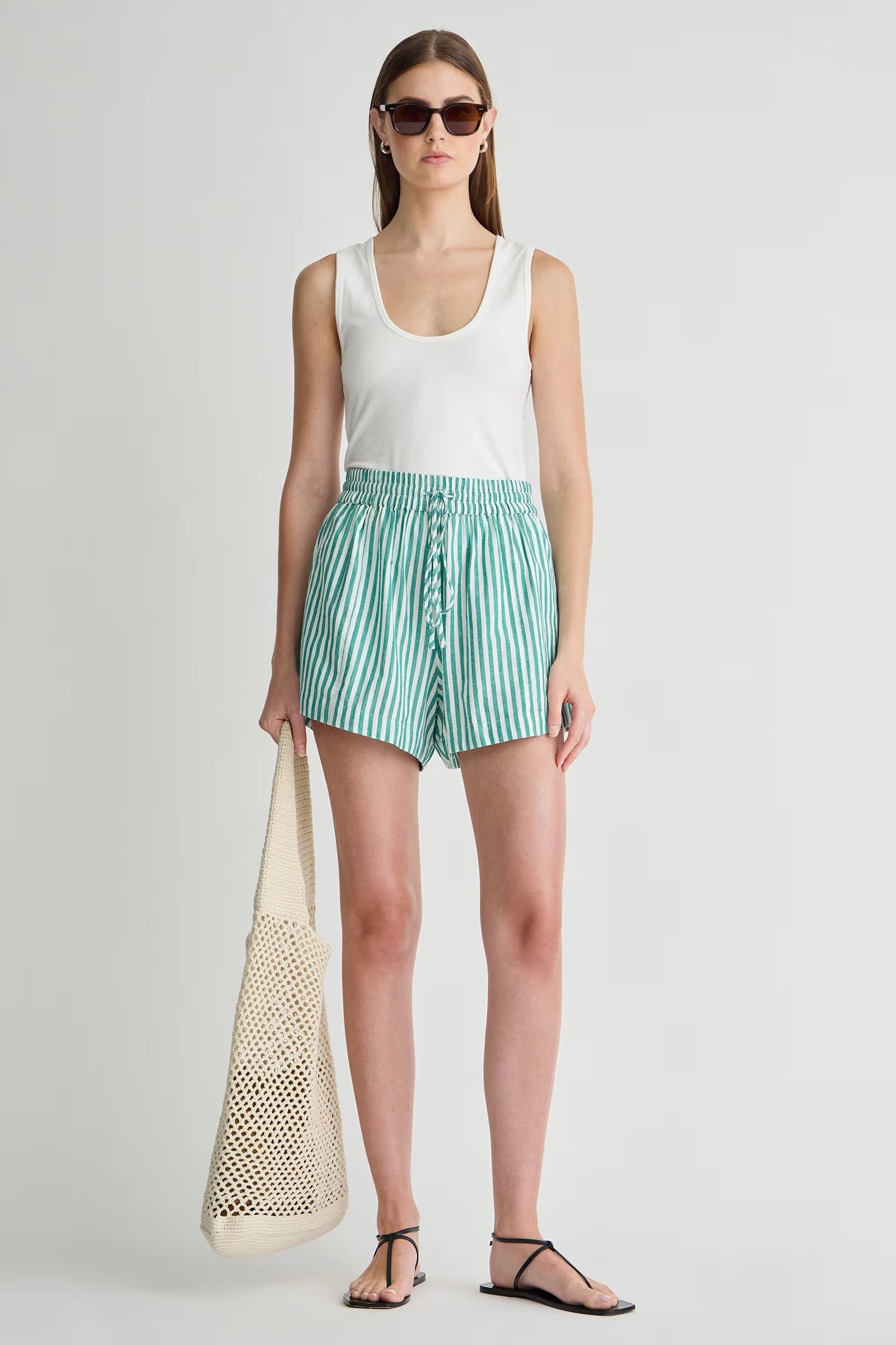 Woman wearing a white sleeveless top and green striped shorts, holding a beige bag on a light gray background.