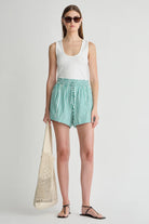Woman wearing a white sleeveless top and green striped shorts, holding a beige bag on a light gray background.