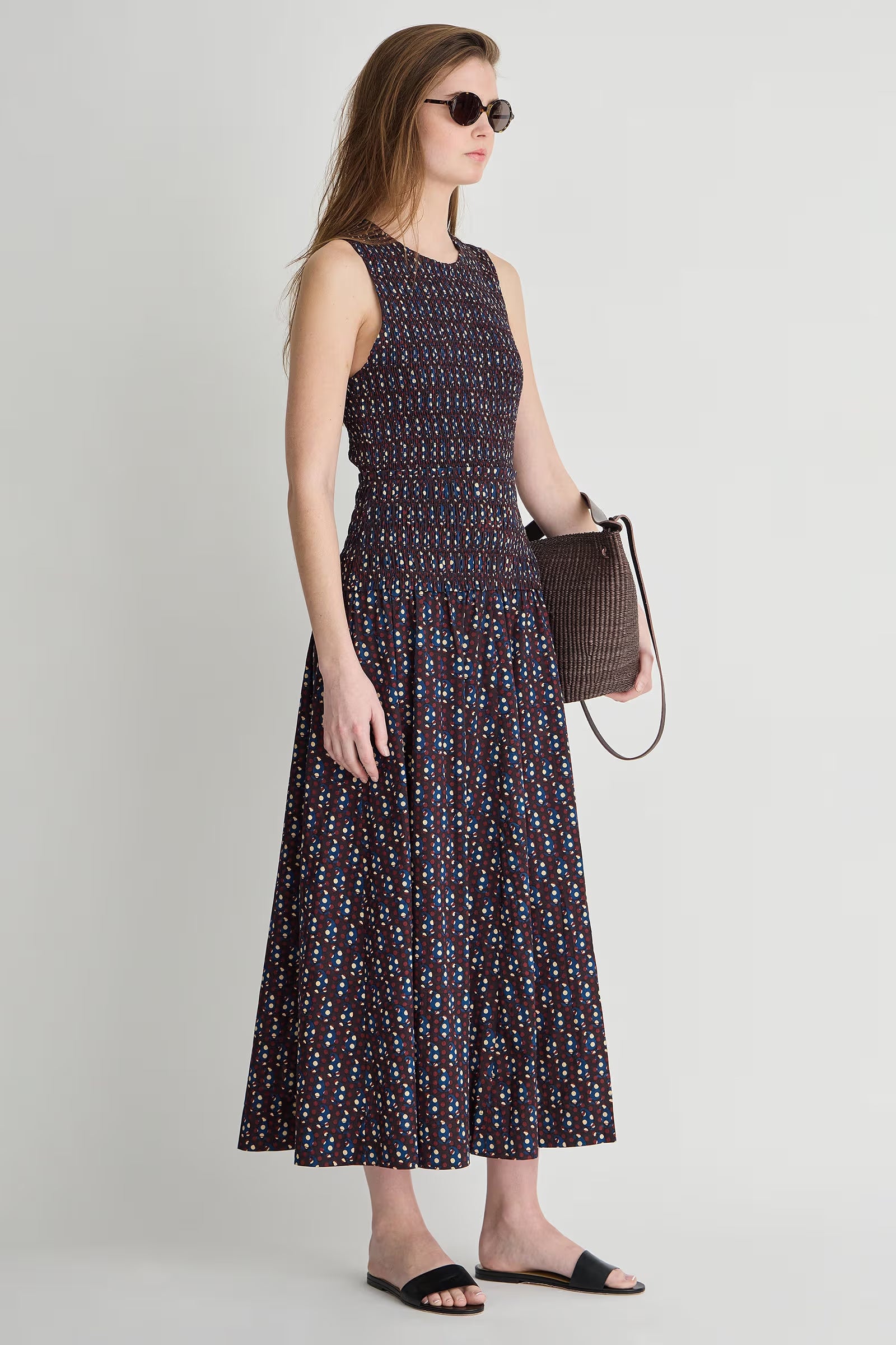 Woman wearing a sleeveless patterned dress holding a brown bag against a plain background
