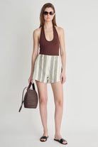 Woman wearing a brown top and striped shorts, holding a woven bag on a white background