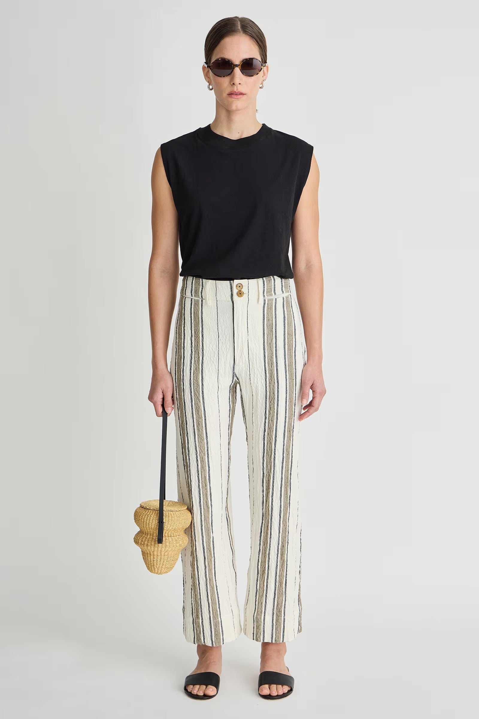 Woman wearing a black sleeveless top and striped pants, holding a woven bag on a light gray background