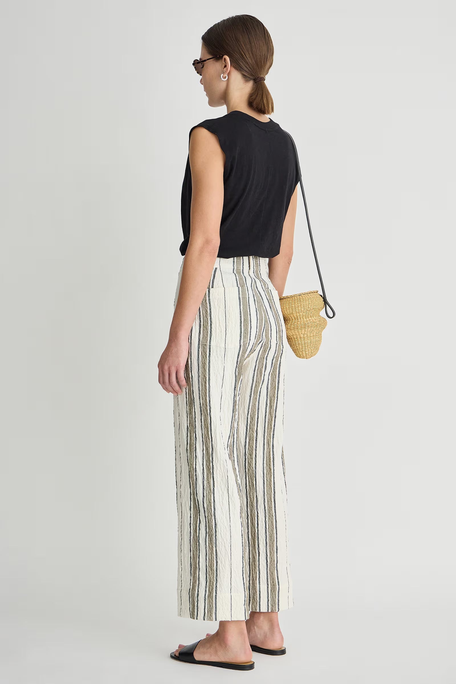 Woman wearing a black sleeveless top and striped pants, holding a straw bag on a white background