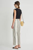 Woman wearing a black sleeveless top and striped pants, holding a straw bag on a white background