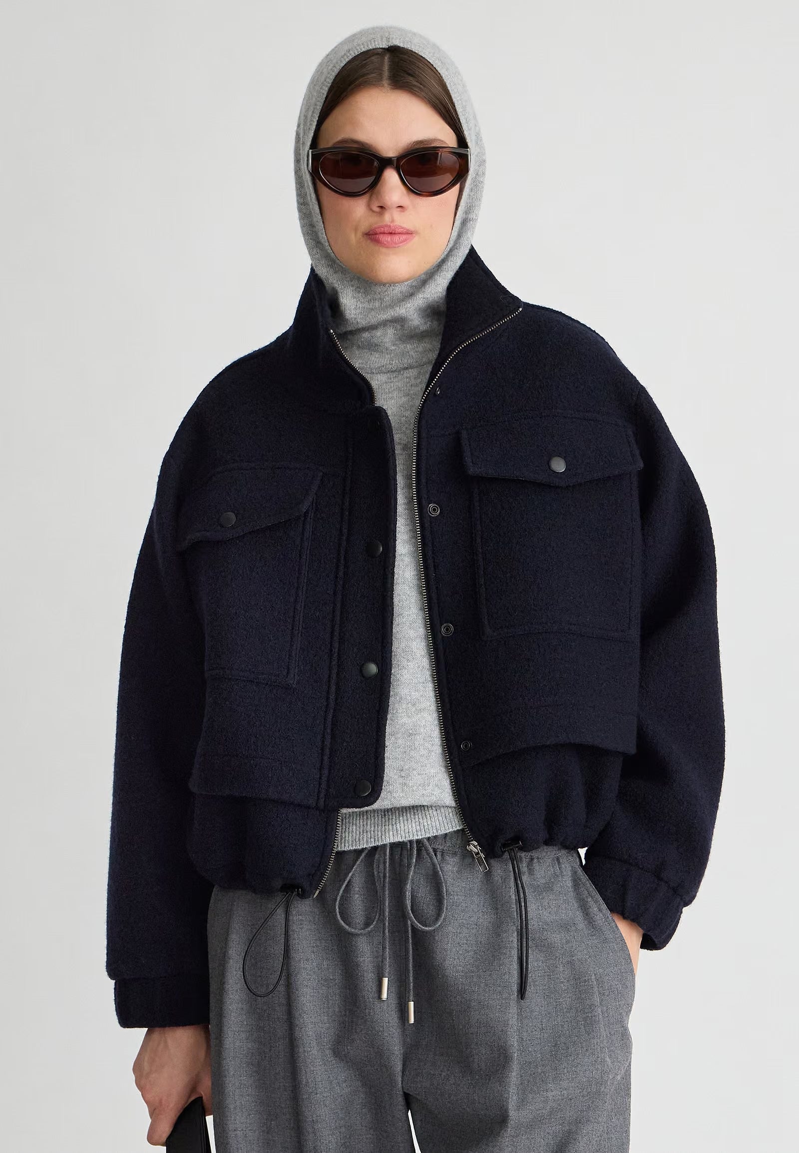 A model wearing a dark navy cropped utility jacket over a gray turtleneck and matching gray drawstring trousers, styled with a gray hood and sunglasses, against a light gray studio background.