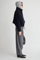 A model shown in profile wearing a dark navy cropped jacket, gray wide-leg trousers, a gray beanie, and black shoes against a light gray studio background.