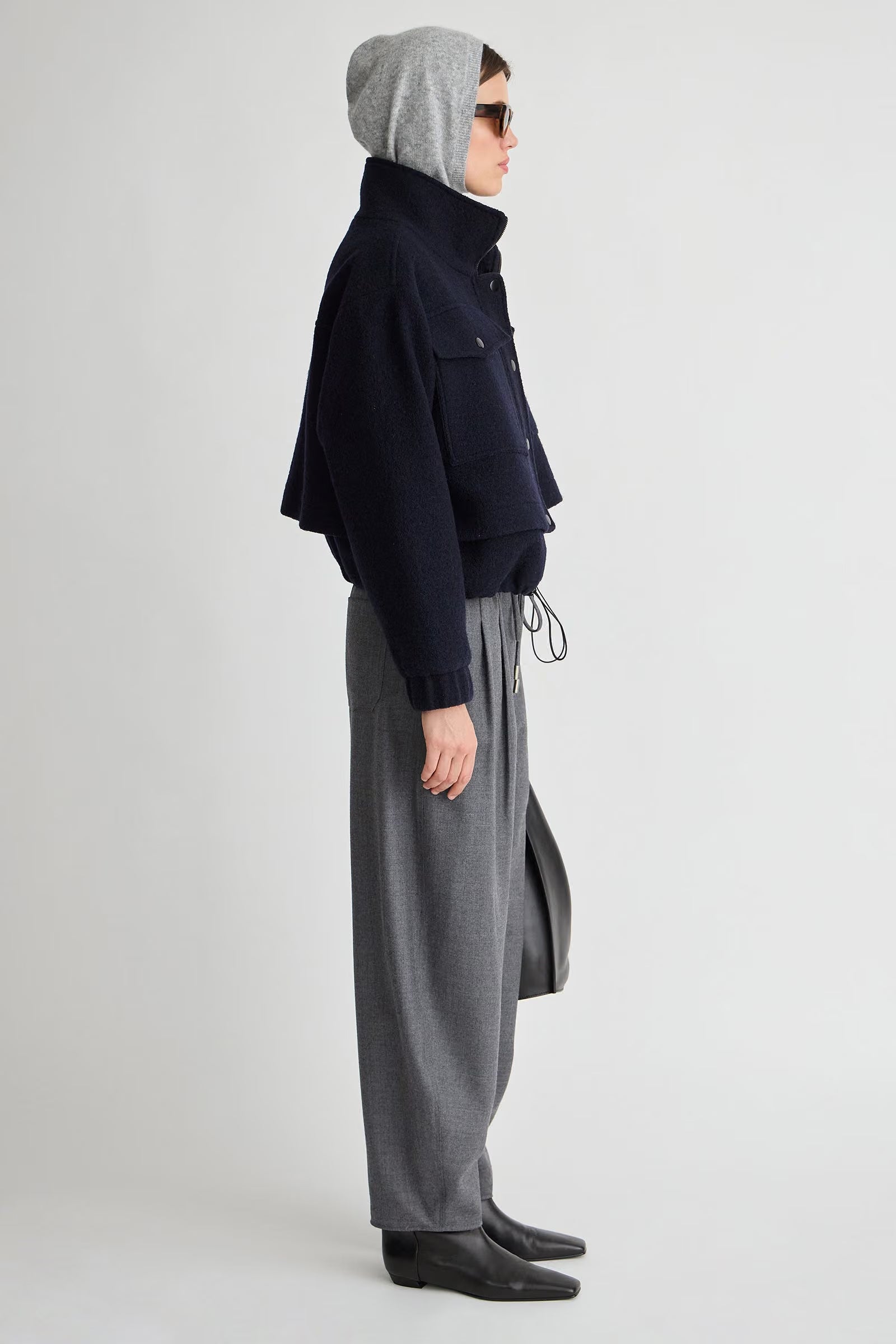 A model shown in profile wearing a dark navy cropped jacket, gray wide-leg trousers, a gray beanie, and black shoes against a light gray studio background.