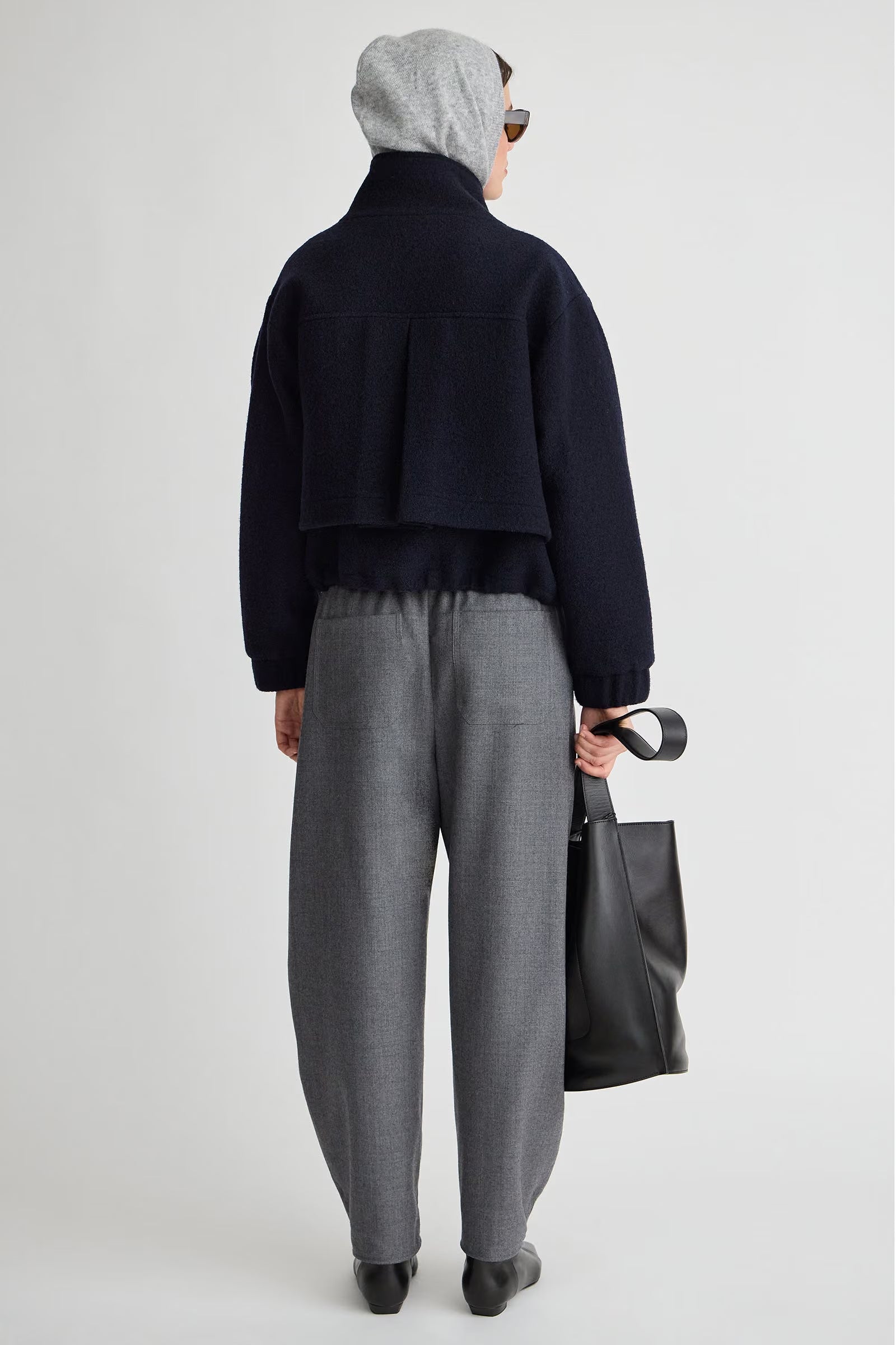 A model shown from the back wearing a dark navy cropped jacket, gray textured trousers, a light gray beanie, and sunglasses, holding a black tote bag against a light gray studio background.