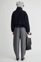 A model shown from the back wearing a dark navy cropped jacket, gray textured trousers, a light gray beanie, and sunglasses, holding a black tote bag against a light gray studio background.