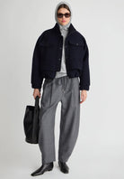 A model stands against a light gray studio background wearing a dark navy cropped jacket over a light gray turtleneck, paired with loose gray drawstring trousers, black pointed boots, a gray head covering, sunglasses, and a black tote bag.