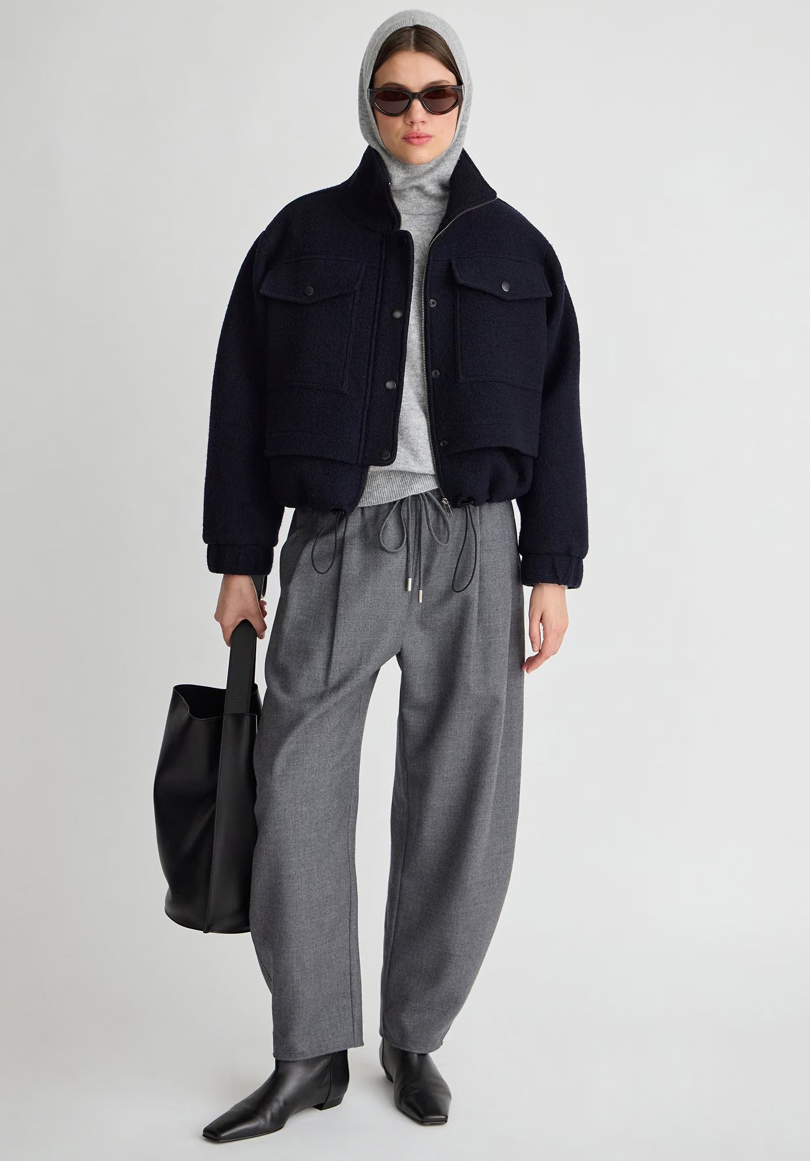 A model stands against a light gray studio background wearing a dark navy cropped jacket over a light gray turtleneck, paired with loose gray drawstring trousers, black pointed boots, a gray head covering, sunglasses, and a black tote bag.
