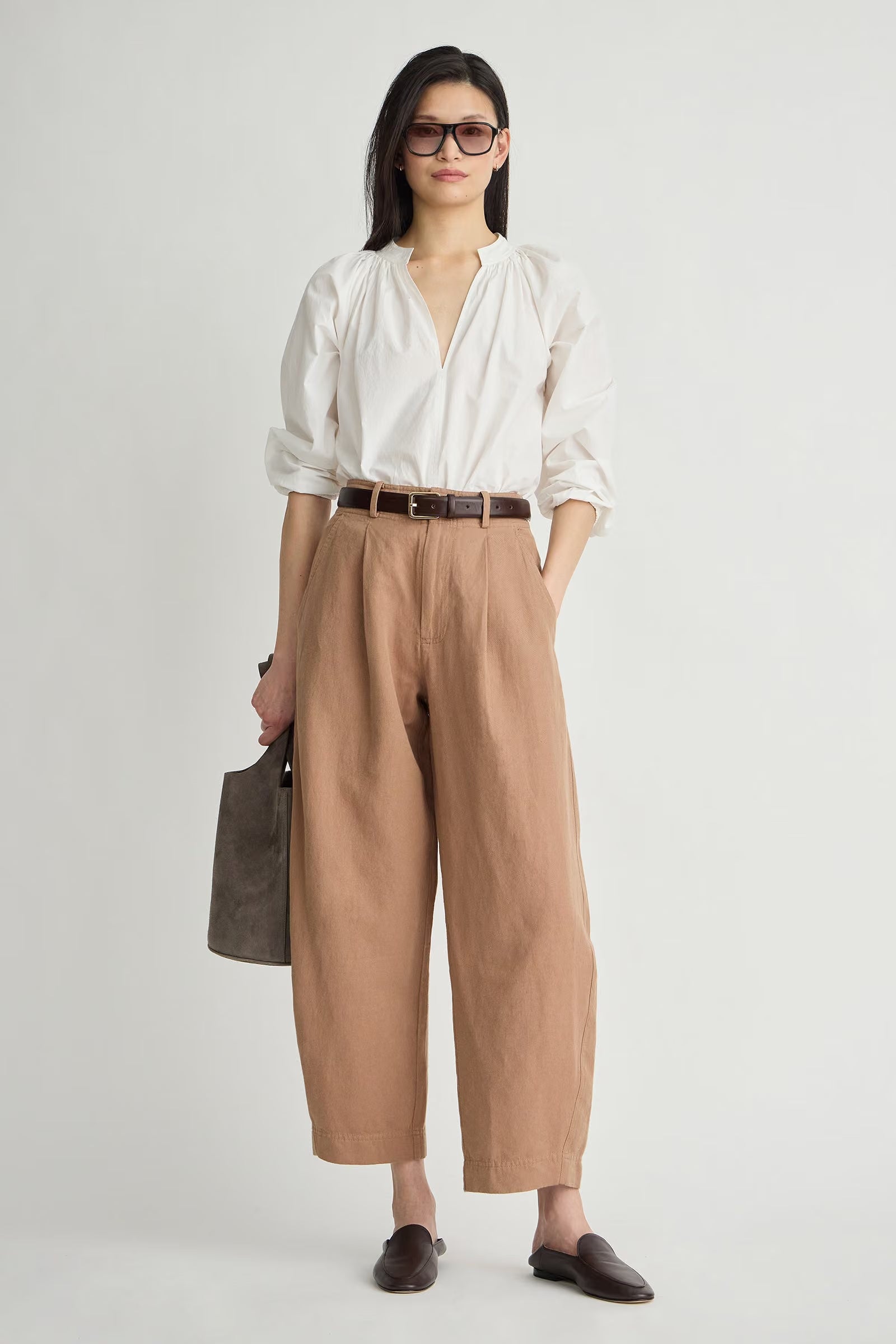 A woman modeling a white long-sleeve blouse tucked into high-waisted, wide-leg tan trousers, styled with a brown belt, dark brown loafers, and a dark gray handbag against a light gray studio background.
