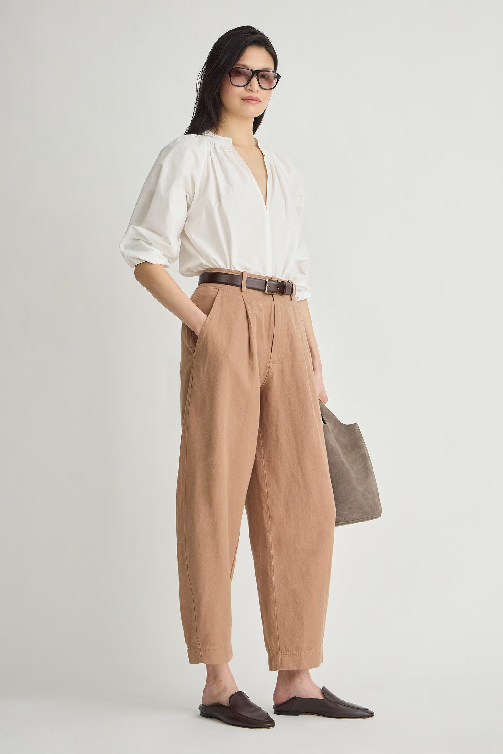 A woman modeling a white blouse and tan wide-leg trousers with a brown belt, standing against a light gray studio background and holding a taupe tote bag.