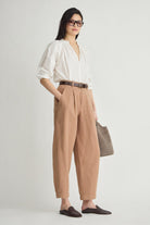 A woman modeling a white blouse and tan wide-leg trousers with a brown belt, standing against a light gray studio background and holding a taupe tote bag.