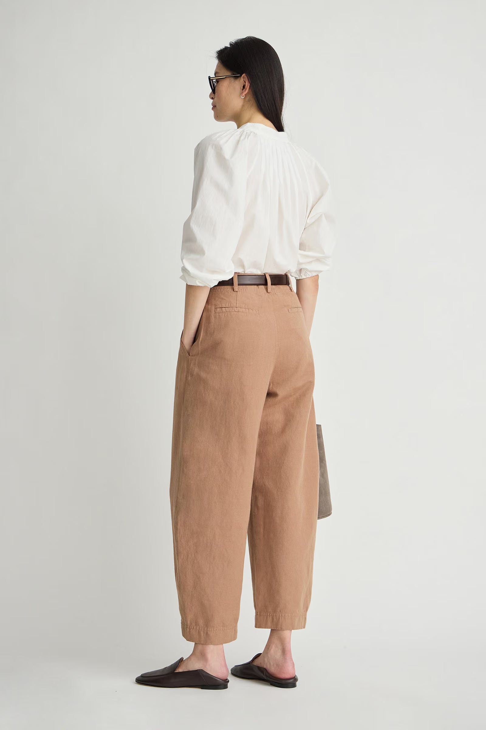 A woman is shown from the back wearing a white blouse, tan wide-leg cropped trousers, a thin brown belt, and black mule sandals against a plain light gray studio background.